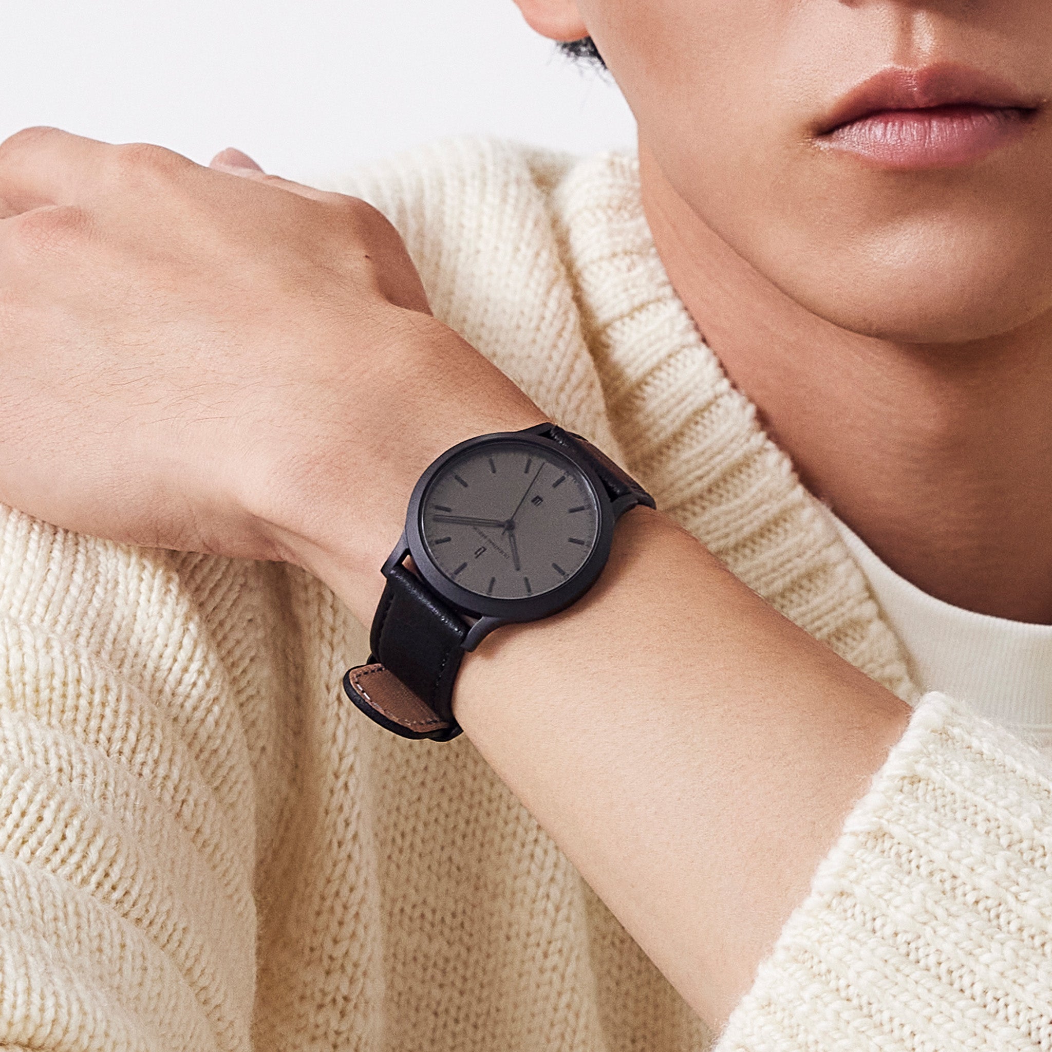 Minimalist black wristwatch with black leather strap on wrist, worn over cream knit sweater