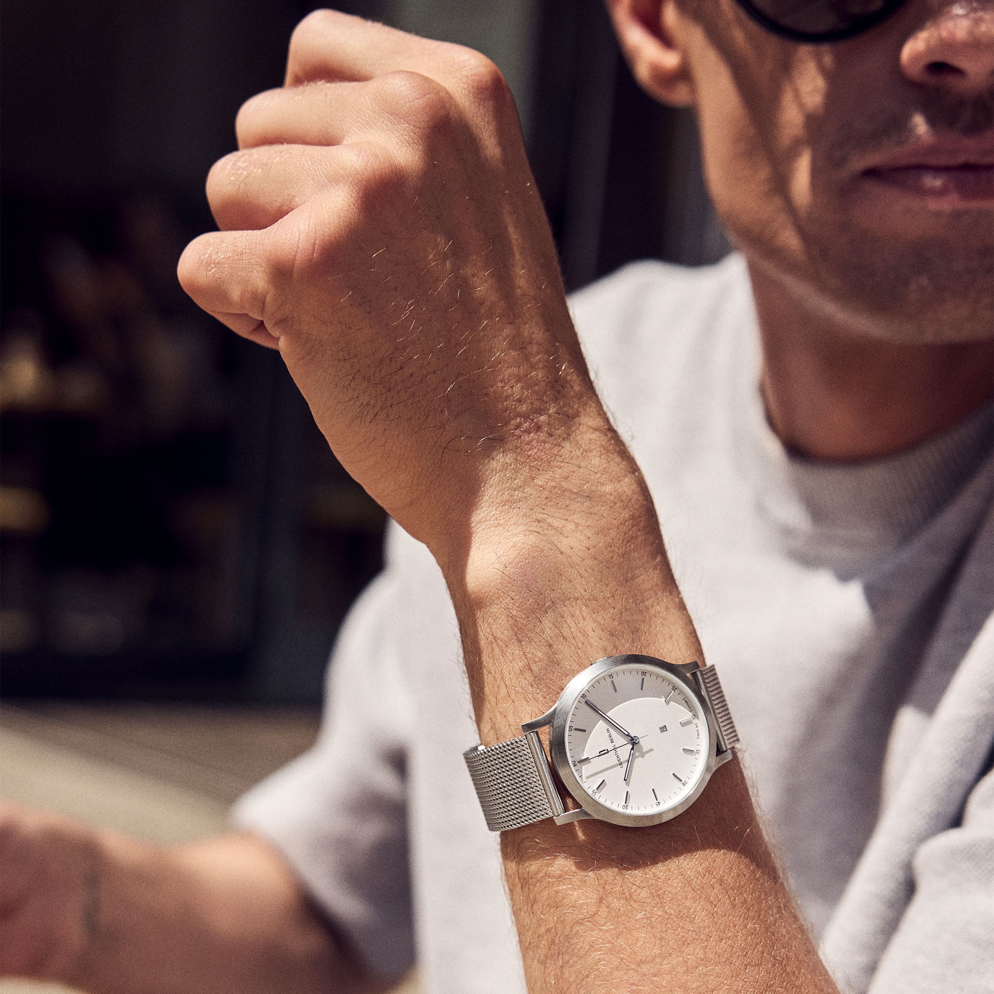 Man wearing a minimalist silver mesh wristwatch with white dial in sunlight