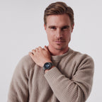 Man in beige sweater showcasing stylish black wristwatch against plain background