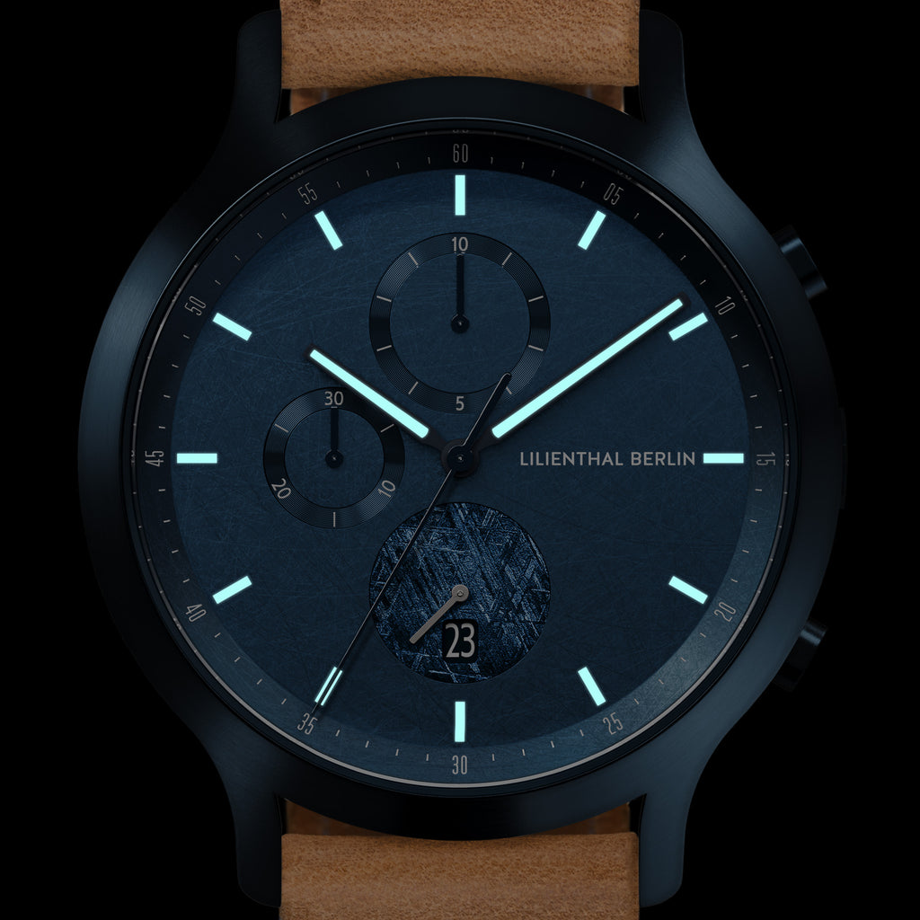 Men's modern black chronograph watch with blue dial, subdials, date window, and tan leather strap.