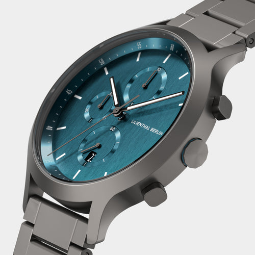 Close-up of a modern chronograph watch with a teal dial, stainless steel strap, and subdials.