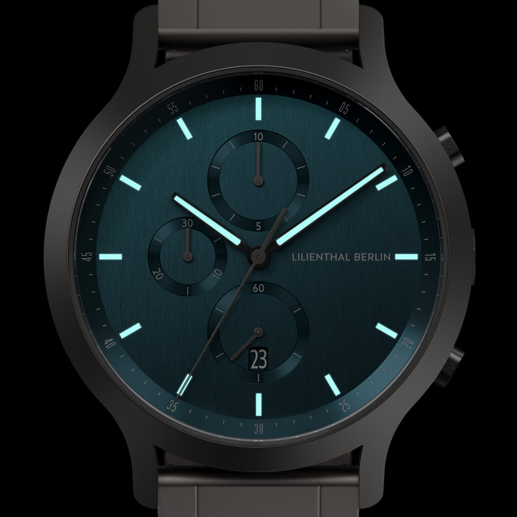 Modern black chronograph watch with teal face, luminous hands, and date display.