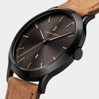 Minimalist black watch with brown dial, gold markers, date display, and tan suede strap