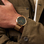 Gold mesh strap wristwatch with green dial on person's wrist, styled with olive jacket