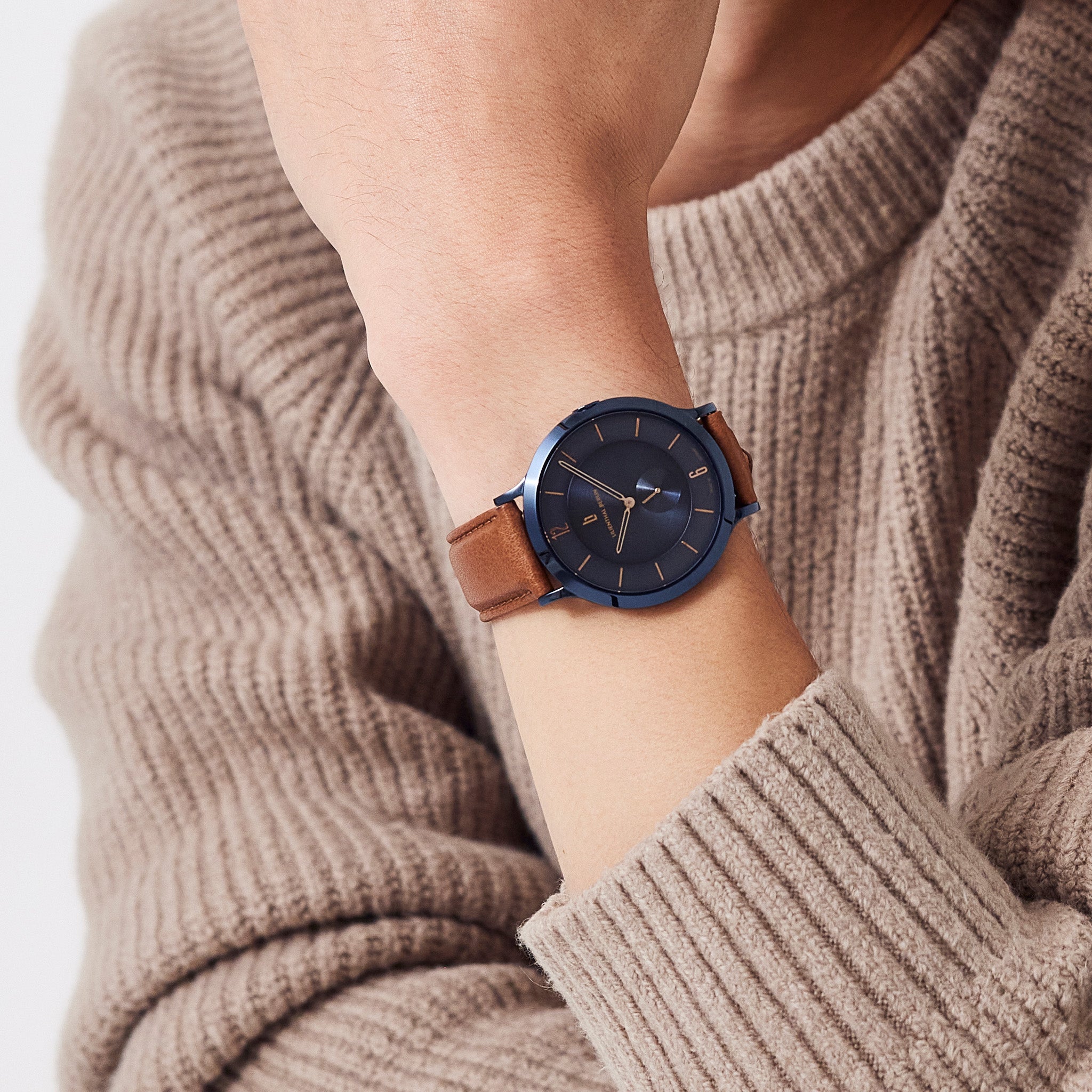 Minimalist blue dial watch with brown leather strap on wrist, worn with beige sweater